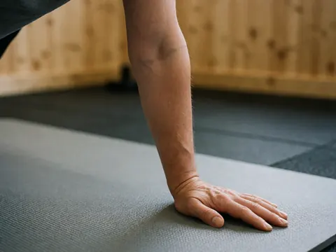 Close-up of an adult hand on a gym mat with a blurred wooden wall and gym floor in the background, highlighting exercise details.