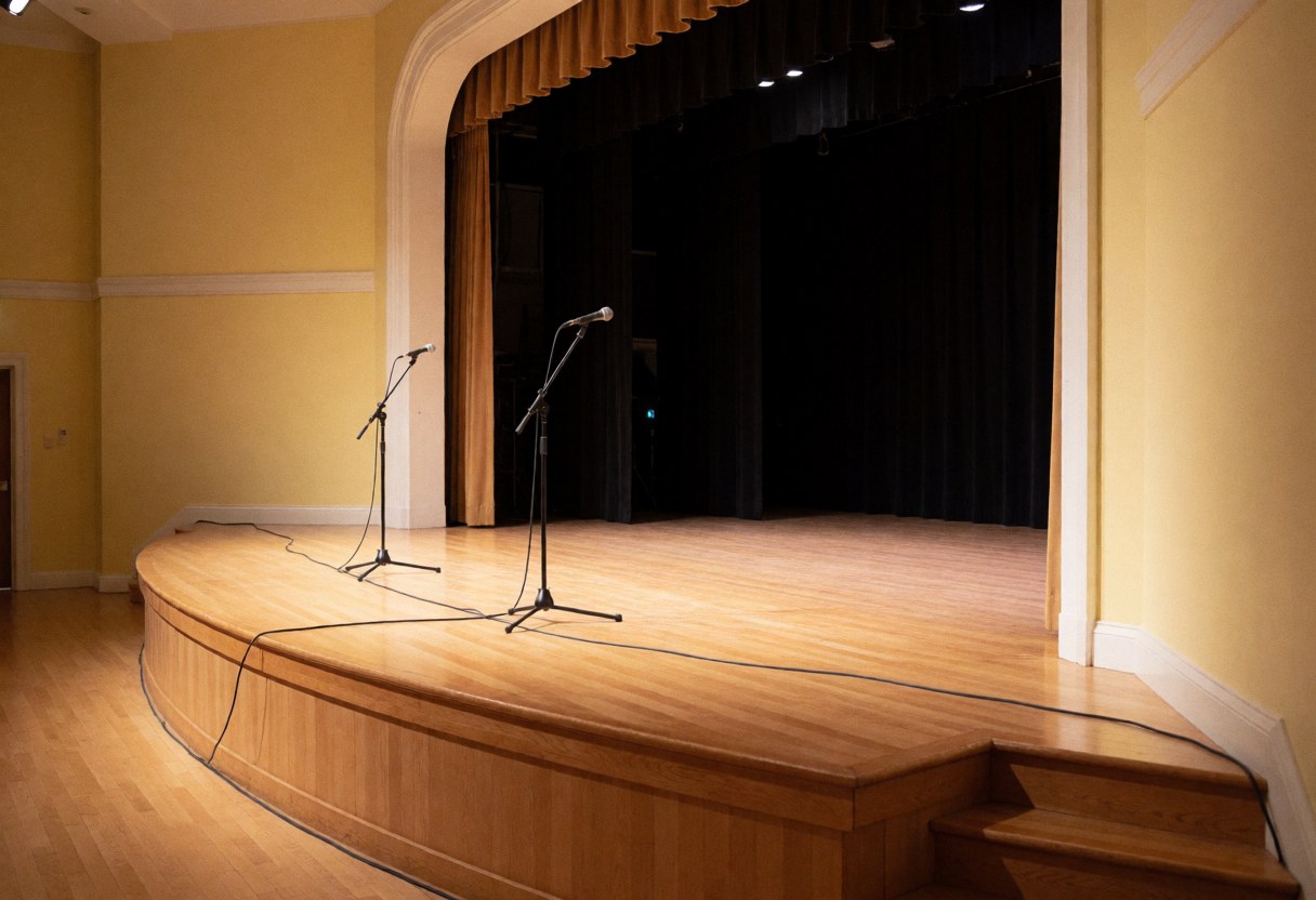 Curved wooden stage with two microphones, cables, black curtains, yellow walls, and warm lighting ready for events