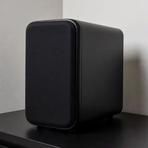 Matte black subwoofer resting on a black table with light gray walls and soft professional lighting