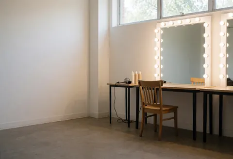 Empty backstage dressing room featuring an LED makeup mirror, wooden chair, hair dryer, and bottles on a table with black legs against white walls and concrete floor