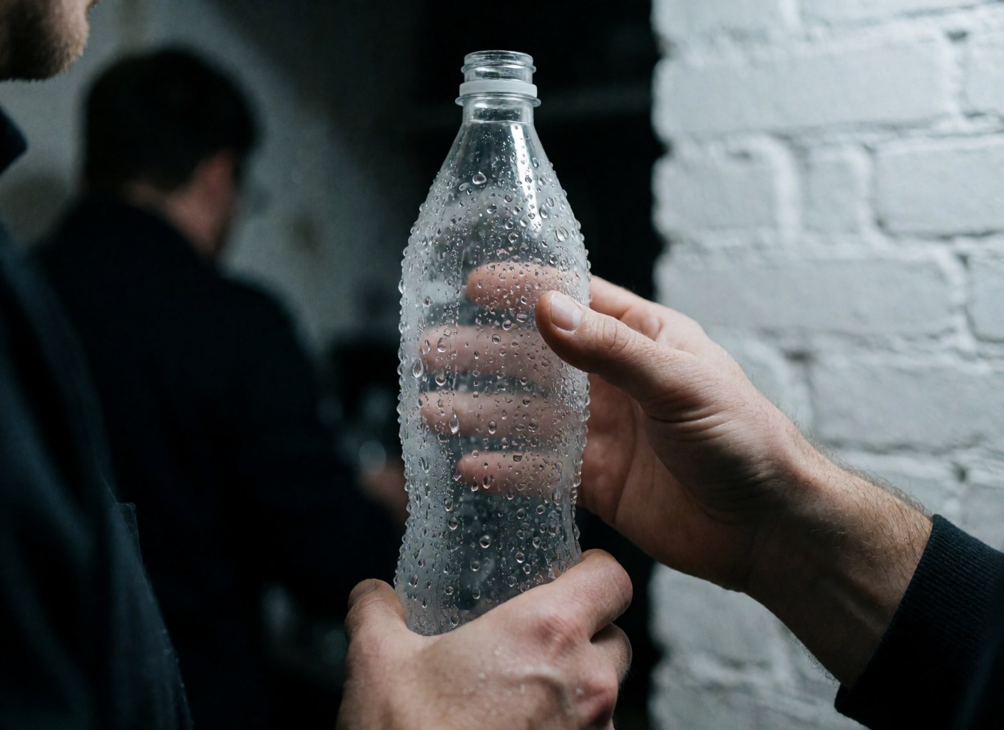 Hands holding a plastic bottle with condensation droplets against a white brick wall background with cool, realistic lighting.