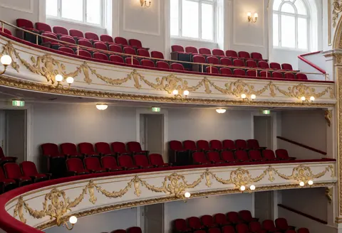 View of empty theater balconies featuring red seating, gold railings, and gray walls illuminated by natural light from high windows.