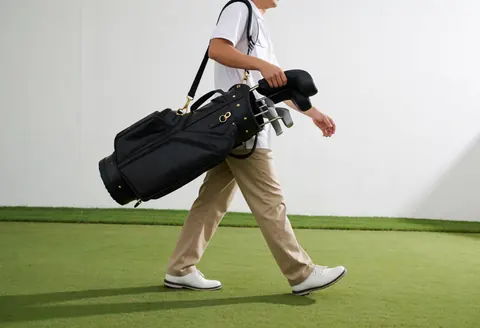 A professional caddie in white uniform and khaki pants walks across the golf course carrying a golf bag under morning light.