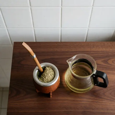 Yerba mate gourd with wooden bombilla and water carafe on dark wood table with white tiled wall and natural light