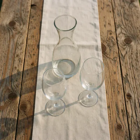 Wine carafe and two wine glasses placed on a rustic wooden table with a linen tablecloth, illuminated by natural light.