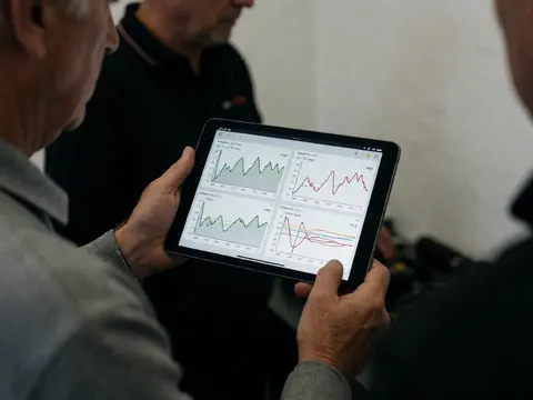 Hands holding a tablet displaying performance graphs at a motorsport circuit control area with a blurred white background