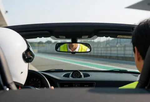 Interior view of a circuit car showing an instructor in a yellow vest reflected in the rearview mirror, ready to drive on the track