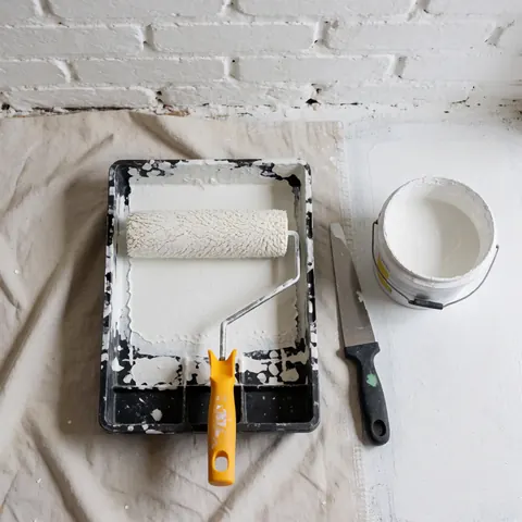 White paint roller in tray, putty knife, and paint bucket on protective cloth next to white brick wall