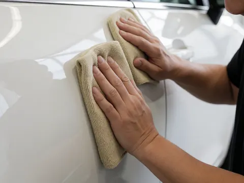 Close-up of a hand using a microfiber cloth to polish the door of a shiny white car, highlighting reflections against a plain white background.