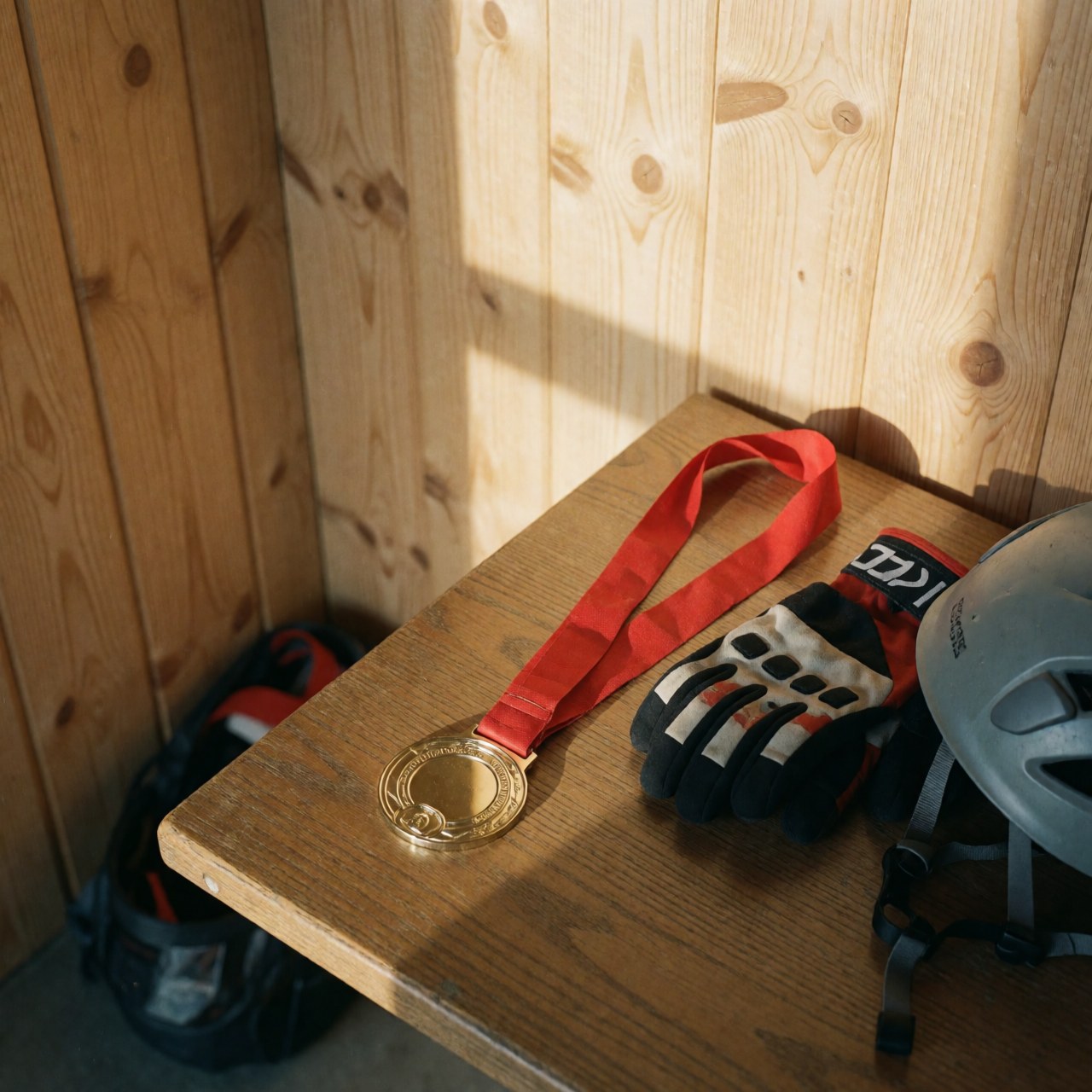 Gold medal with red ribbon, climbing gloves, and helmet on wooden table with wood panel walls