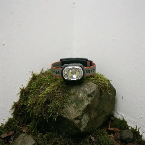 A modern headlamp placed on a wet moss-covered rock against a white background, suitable for outdoor night adventures.