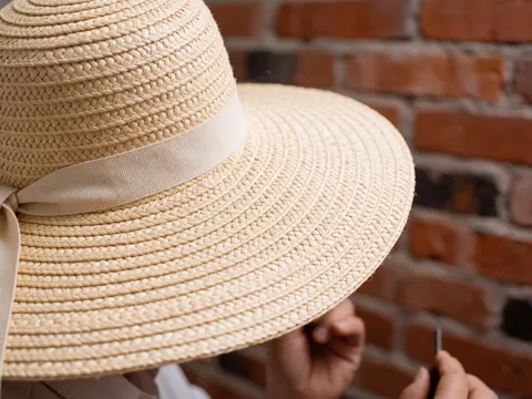 Close-up of a straw hat with a beige ribbon against a red brick wall background.