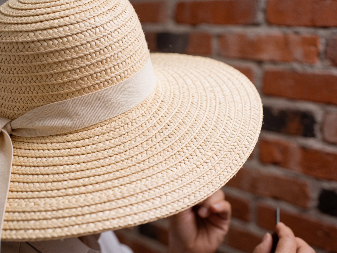Close-up of a straw hat with a beige ribbon against a red brick wall background.