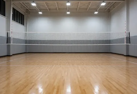 Empty indoor volleyball court featuring polished wooden flooring, a central net, and gray walls with safety zones, ideal for training.