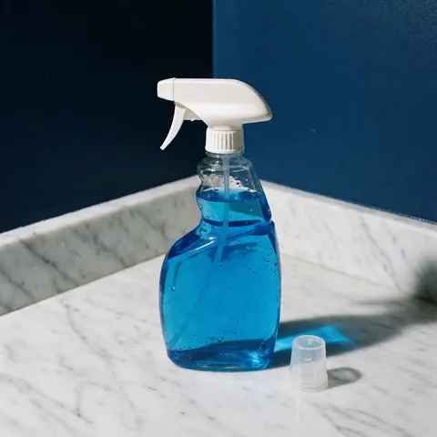 Shiny blue spray bottle with cap beside it on white marble, dark blue wall background