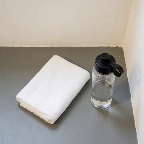 Folded white towel and water bottle with black cap on gray gym floor against warm white walls with soft overhead lighting