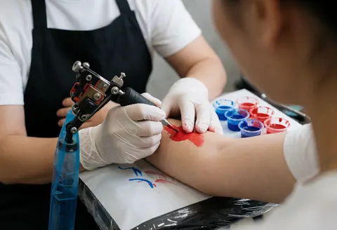 Close-up of a tattoo artist wearing gloves using a machine to apply red ink on a client's arm, with a tray of blue and red inks nearby in a clean studio.