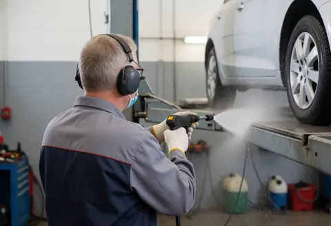 Worker in automotive shop using pressure washer with safety gear to clean a car, surrounded by tools and equipment.