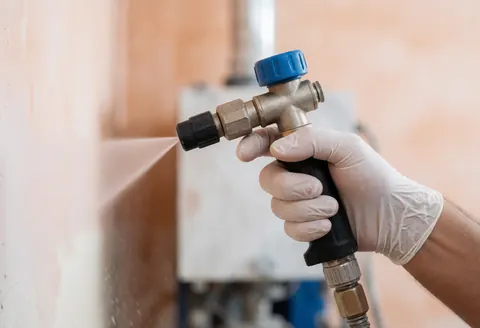 A gloved hand directs an industrial hose spraying water onto a terracotta wall with blurred equipment in the background.