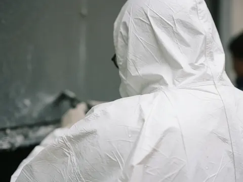 Close-up of a white Tyvek suit shoulder closure featuring visible stitching and water droplets against a blurred industrial background.