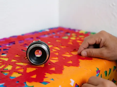 Close-up of a vintage camera lens resting on colorful papel picado paper with sharp details and a white background