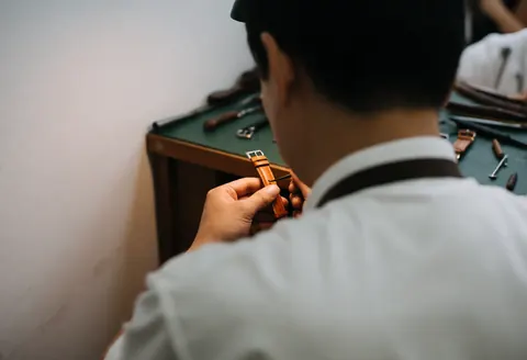 A watchmaker in a white shirt applies treatment to a brown leather strap on a green workbench with tools.