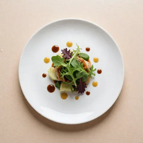 Top-down view of a white plate holding a green salad with brown and yellow sauces on a soft beige background