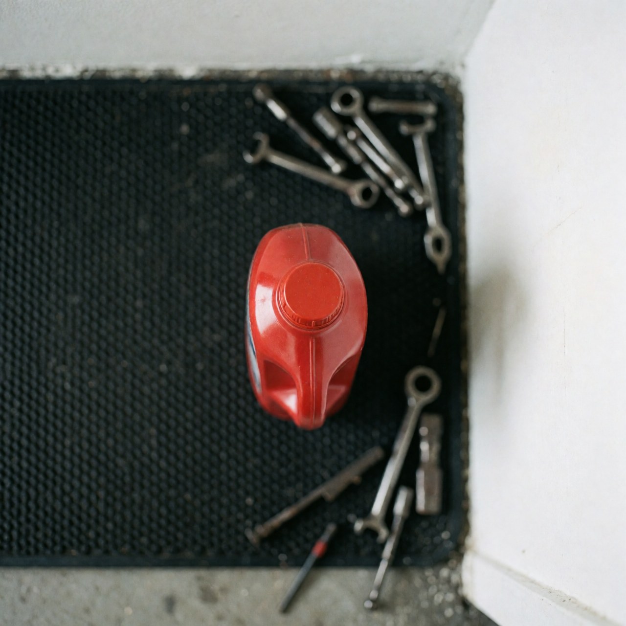 Top view of a red oil bottle on a black mat surrounded by scattered wrenches in a mechanic shop setting