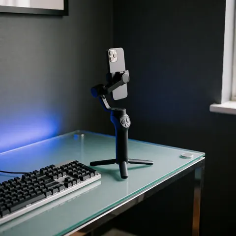 Black gimbal holding a smartphone on a glass desk next to a keyboard under blue light with gray walls