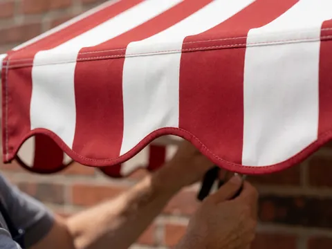 Close-up of hands adjusting the scalloped edge of a red and white awning against a blurred brick wall background with natural lighting
