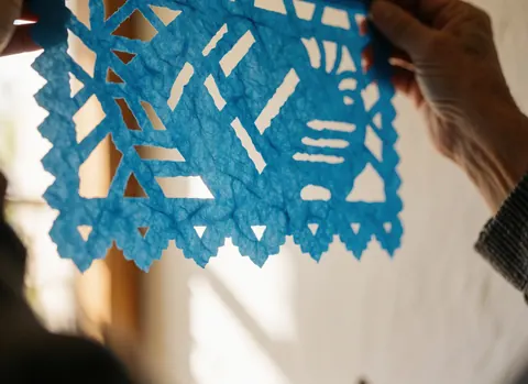 Close-up of hands holding a blue paper flag with sunlight filtering through the cutouts, showing visible paper texture