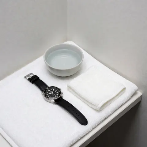 Dry wristwatch with black strap on white towel next to water cup and cloth on light gray corner with bright clean lighting