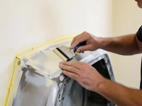 Close-up of a hand using a spatula to apply body filler to a metal car surface, with yellow tape and a white wall in the background