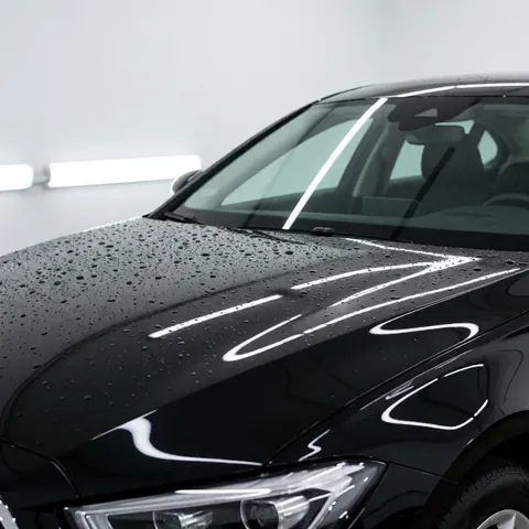 Close-up of a black car hood with water droplets reflecting LED lights in an auto detailing workshop with white walls.
