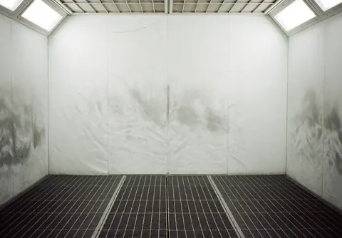 Empty automotive paint booth with white walls and a metal grid floor, illuminated by overhead lights.