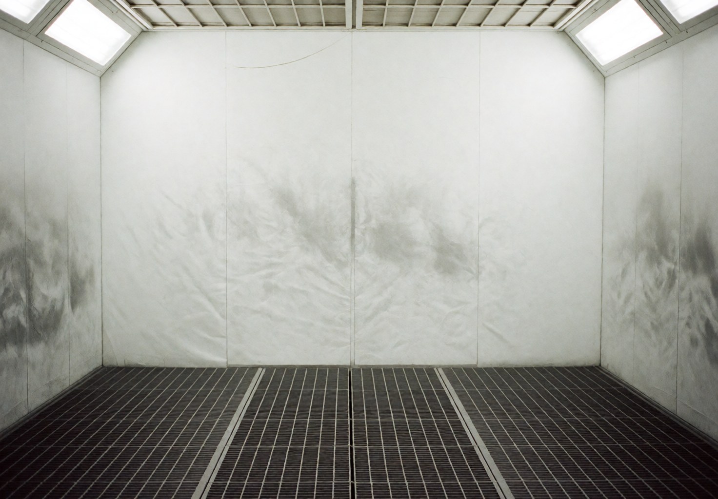 Empty automotive paint booth with white walls and a metal grid floor, illuminated by overhead lights.