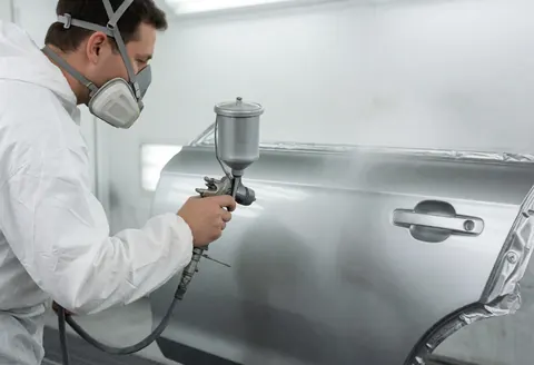 A painter in protective gear applies metallic paint to a car door in a professional auto body shop with bright, even lighting.