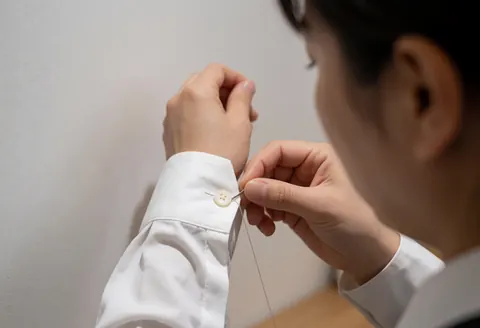 Adult hand stitching a button onto the sleeve of a white shirt against a white background with natural light