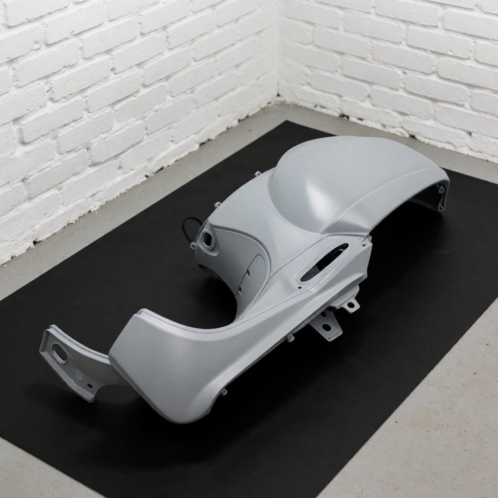 Gray painted scooter body panel resting on a black mat with a white brick wall background and professional lighting