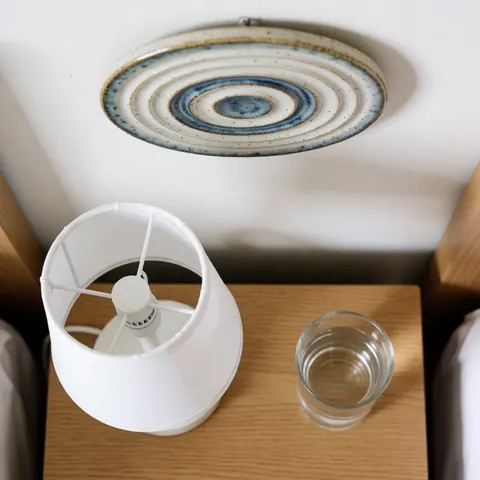 Minimalist wooden nightstand featuring a white lamp, a glass of water, and ceramic wall art in a clean, cozy bedroom setting.