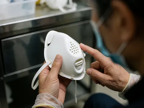 A professional wearing gloves inspects a white medical mask with ventilation holes in a clinical setting with a metallic background.
