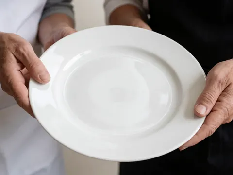 Close-up of adult hands holding a white ceramic plate against a neutral background with bright lighting, suitable for banquet and catering services