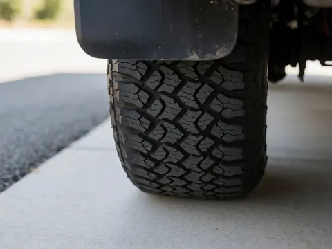 Close-up view of a truck tire tread pattern on concrete with blurred asphalt background and natural light highlighting the rubber texture.