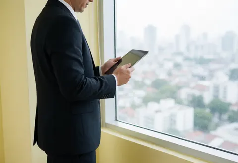 A professional man in a dark suit holds a tablet while looking out a window at a city view against a pale yellow wall.
