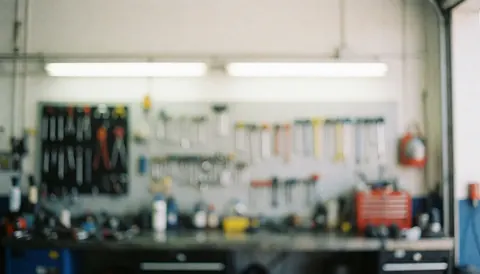 Out of focus auto repair shop background featuring wall-mounted tools, work tables, and fluorescent lighting, suitable for text overlay.