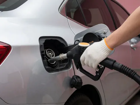 Close-up of a gloved hand inserting a fuel nozzle into the open fuel port of a silver car, revealing the internal mechanism against a pink wall background.