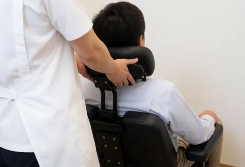 Shiatsu practitioner in white apron applying neck massage to client seated in black ergonomic chair against white wall