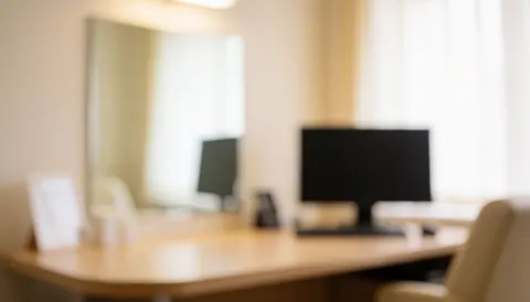 Soft focus medical consultation room featuring a desk, computer, mirror, and chair with warm lighting, suitable for text overlay