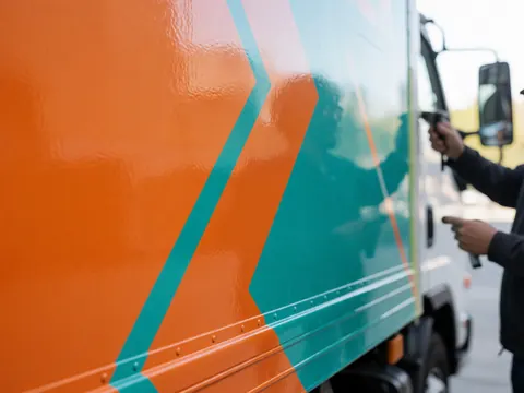 Close-up of hands applying bright orange and turquoise vinyl to a marketing truck side panel under natural light against a plain concrete wall