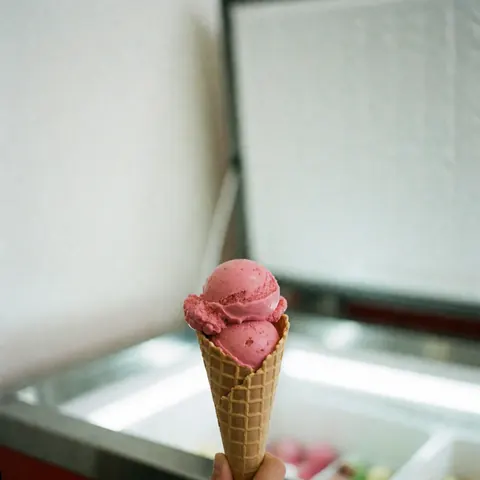A pink strawberry ice cream cone held in front of a bright ice cream shop display case with a white background.